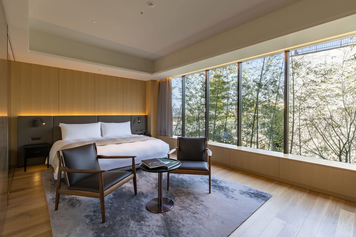 Une chambre moderne avec un grand lit, deux fauteuils, une petite table et une vue sur les arbres à travers de grandes fenêtres.