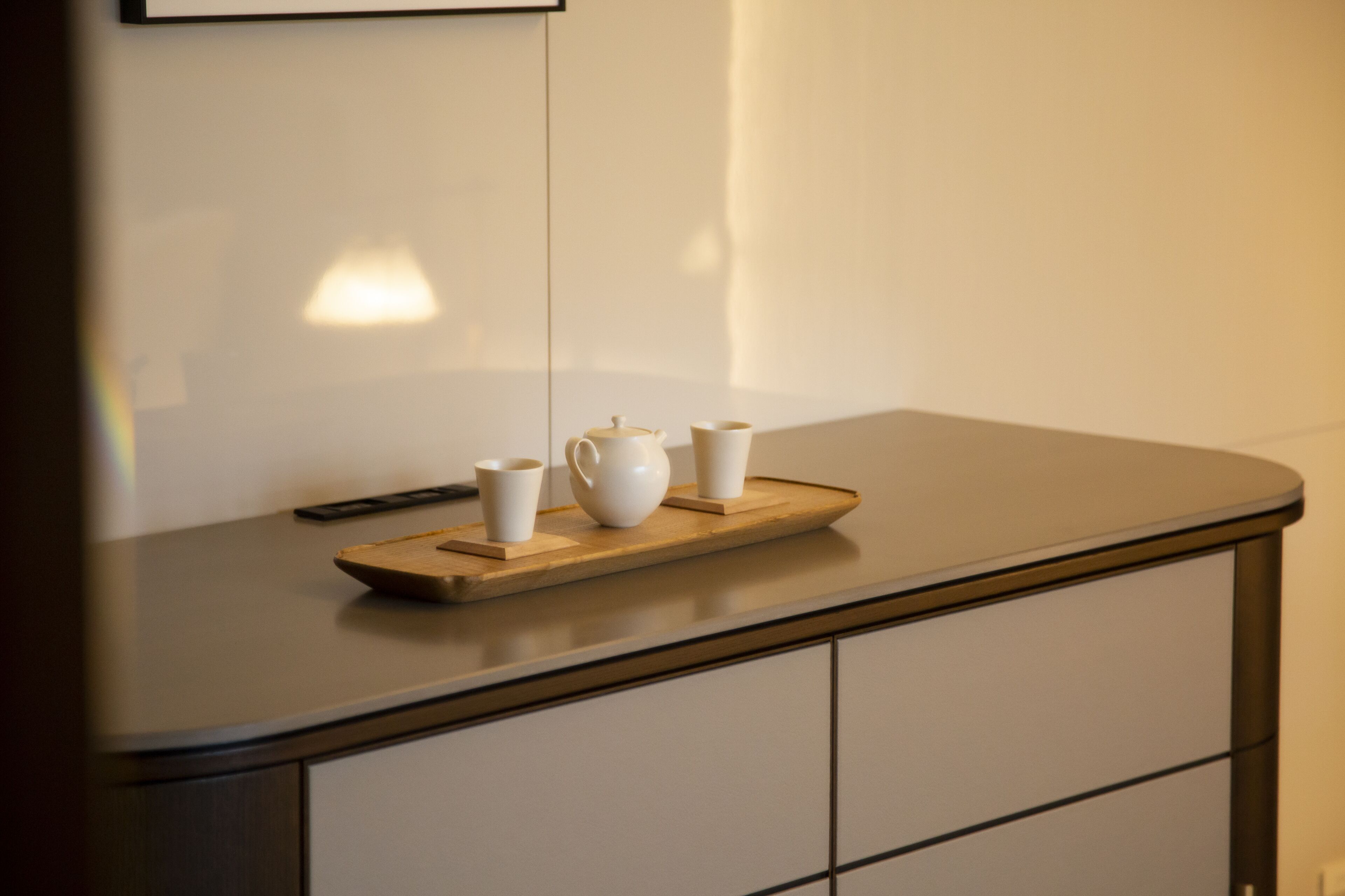 A kitchen counter with a wooden tray holding a teapot and two cups, and a remote control on the left.