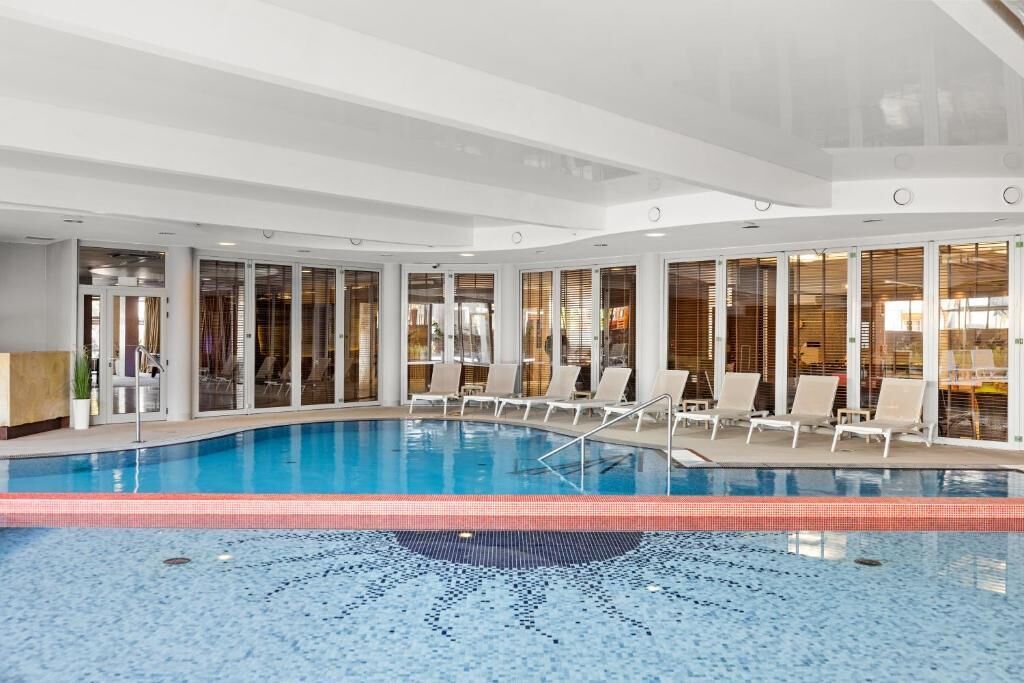 Una piscina interior con paredes de vidrio transparente, sillones de descanso y suelo de baldosas.