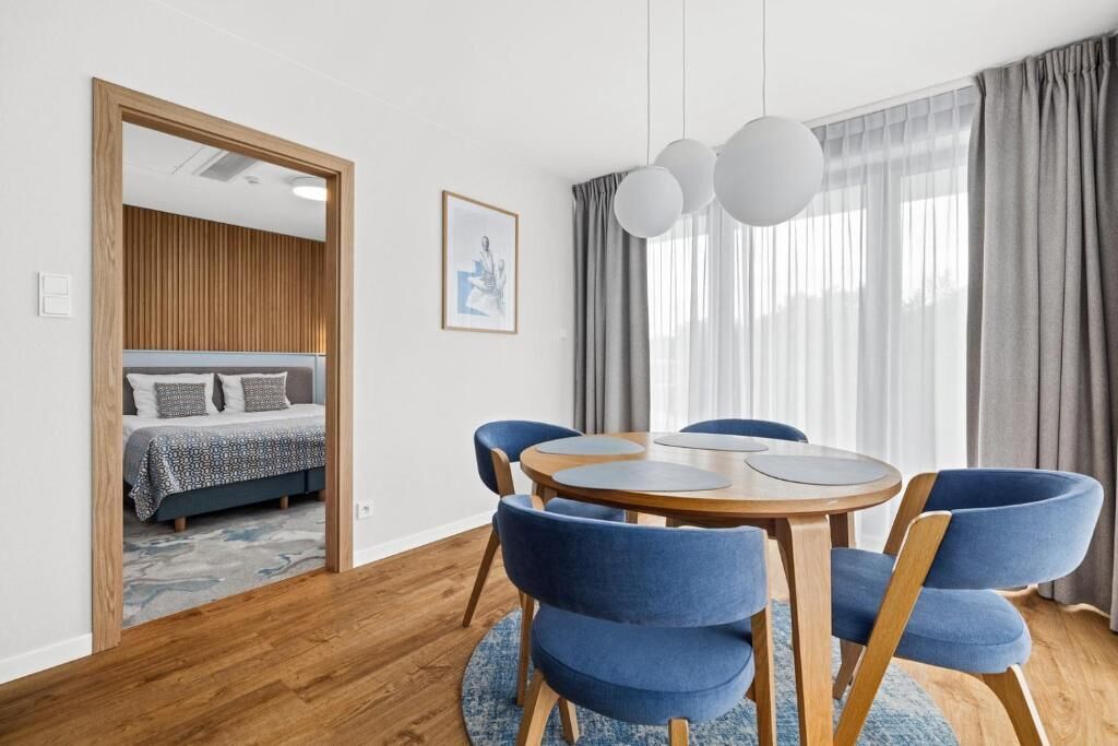 A modern room with a round wooden table, blue upholstered chairs, a bed with patterned bedding, a framed picture on the wall, and a large window with curtains.