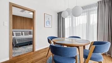 A modern room with a round wooden table, blue upholstered chairs, a bed with patterned bedding, a framed picture on the wall, and a large window with curtains.