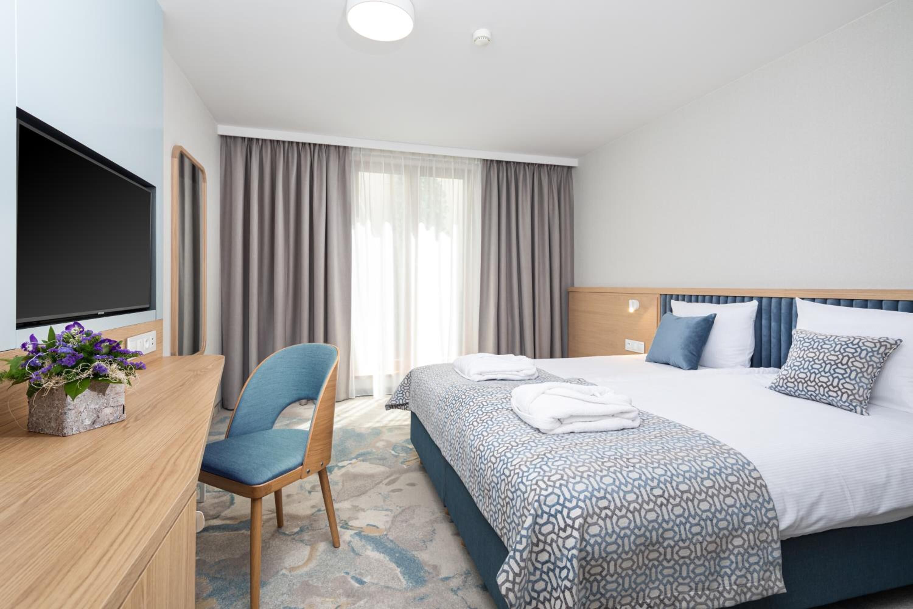 A hotel room with a bed, a desk with a flat-screen TV, a chair, and a window with curtains.