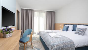 A hotel room with a bed, a desk with a flat-screen TV, a chair, and a window with curtains.