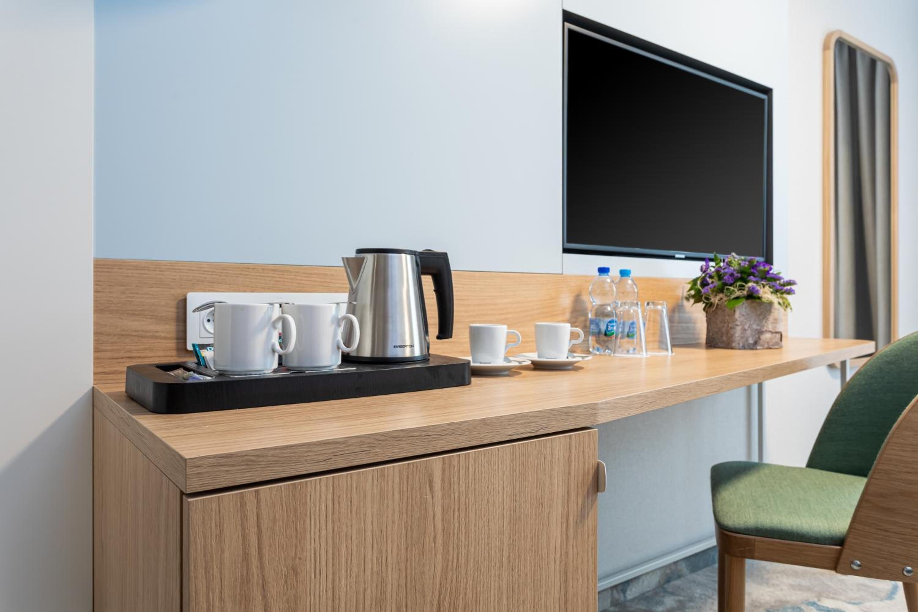 Hotelkamer met een houten bureau, een koffiezetapparaat, kopjes, een waterfles en een bloevas.