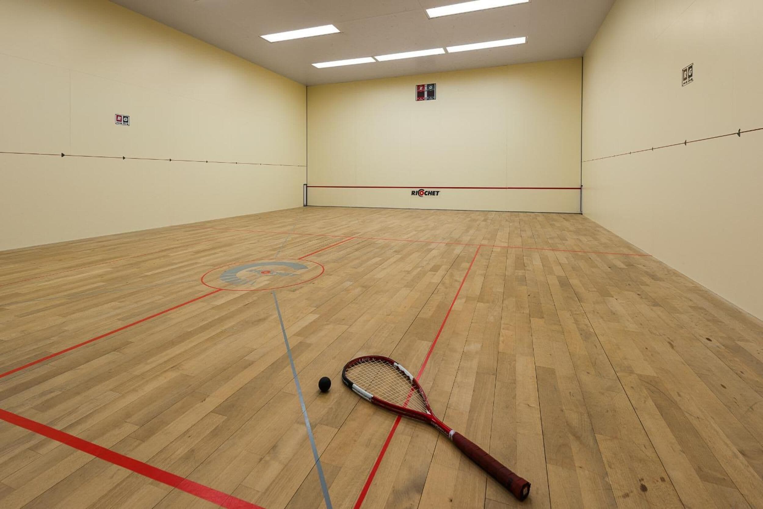 Una cancha de squash con piso de madera, una raqueta roja y negra, y una línea divisoria azul y roja.