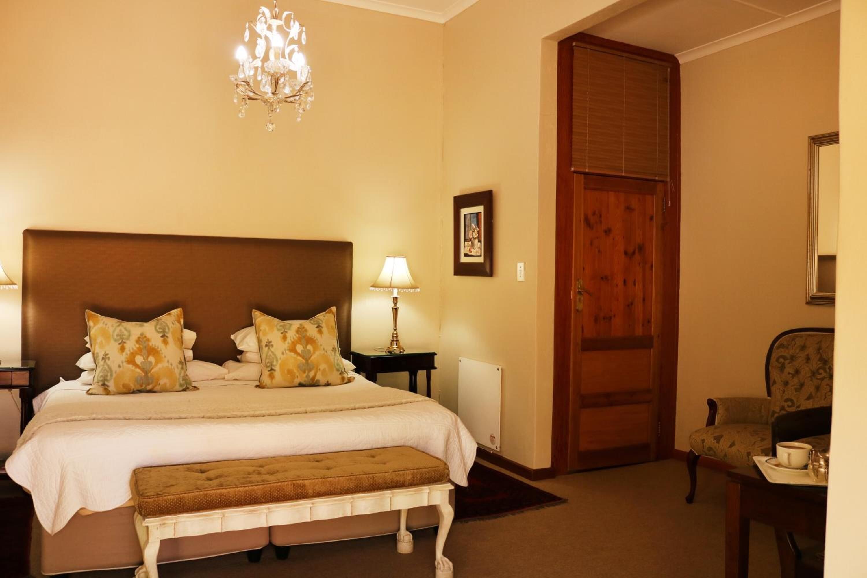 Une chambre d’hôtel avec un grand lit, un banc, un lustre, une porte en bois et une chaise.