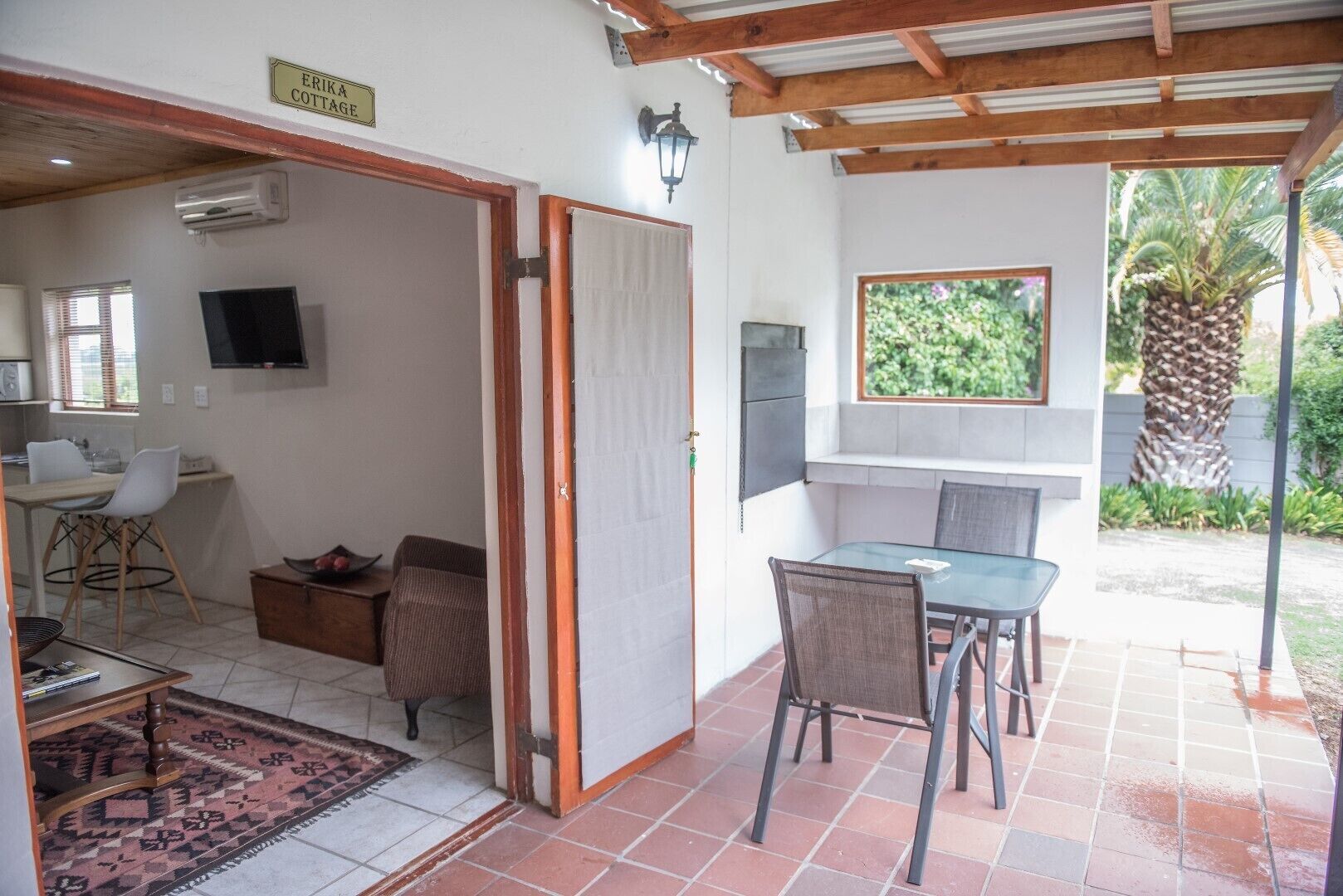 A cozy outdoor patio area with a dining table and chairs, a TV mounted on the wall, and a sign indicating 'ERIKA COTTAGE'.