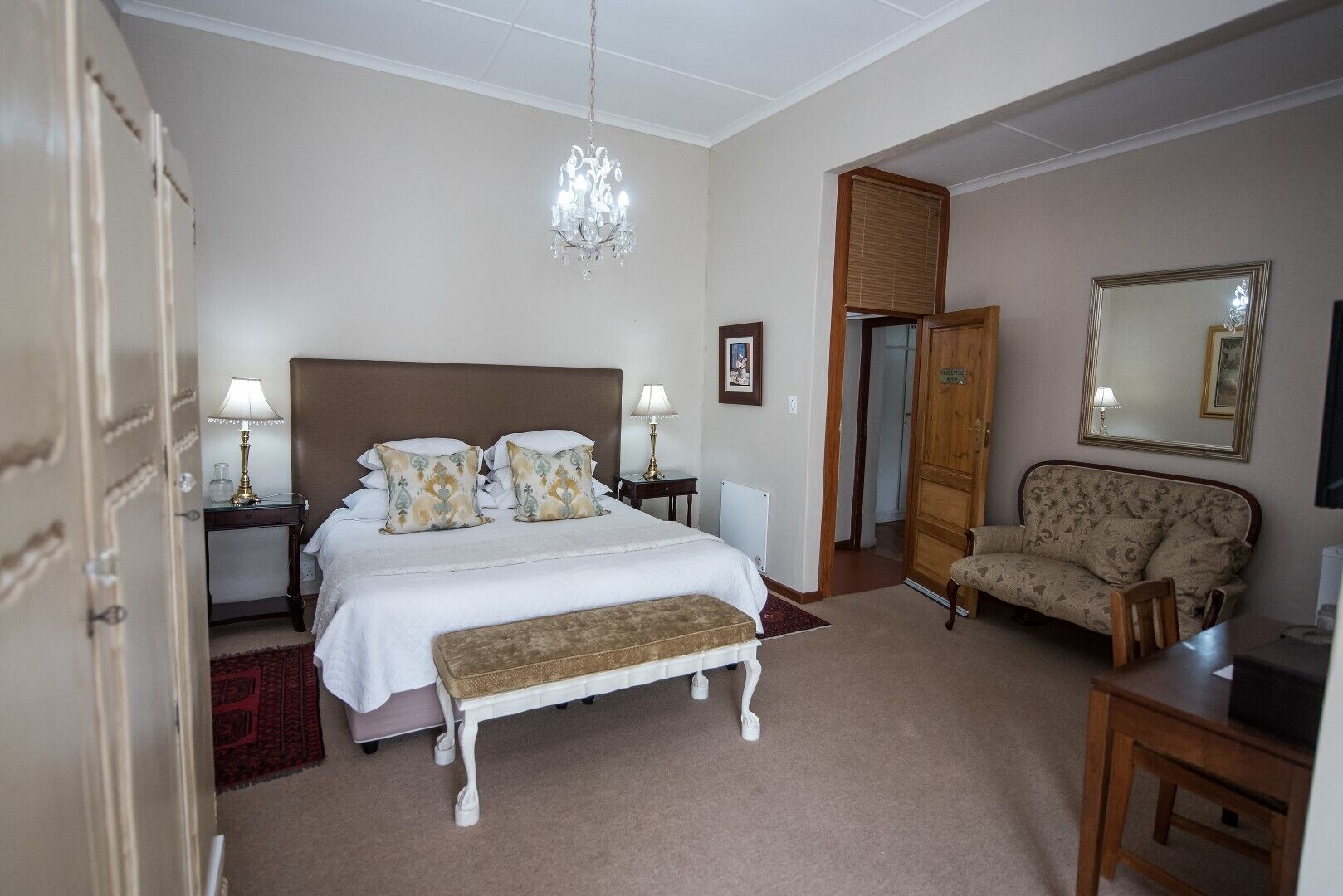 A bedroom with a large bed, bedside tables, a chair, a bench, a chandelier, a mirror, and a wardrobe.