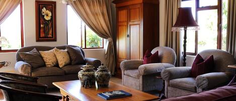 A cozy living room with a sofa, armchairs, a wooden coffee table, and a lamp. There are windows with curtains, a painting on the wall, and a chandelier.