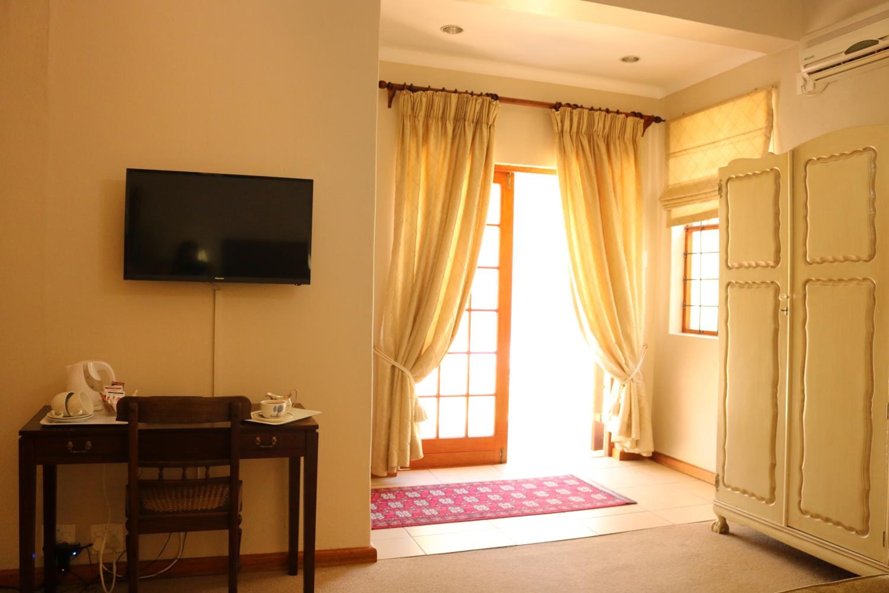 A room with a flat-screen TV mounted on the wall, a wooden desk with a chair, a wardrobe, and a door leading to an outdoor area.