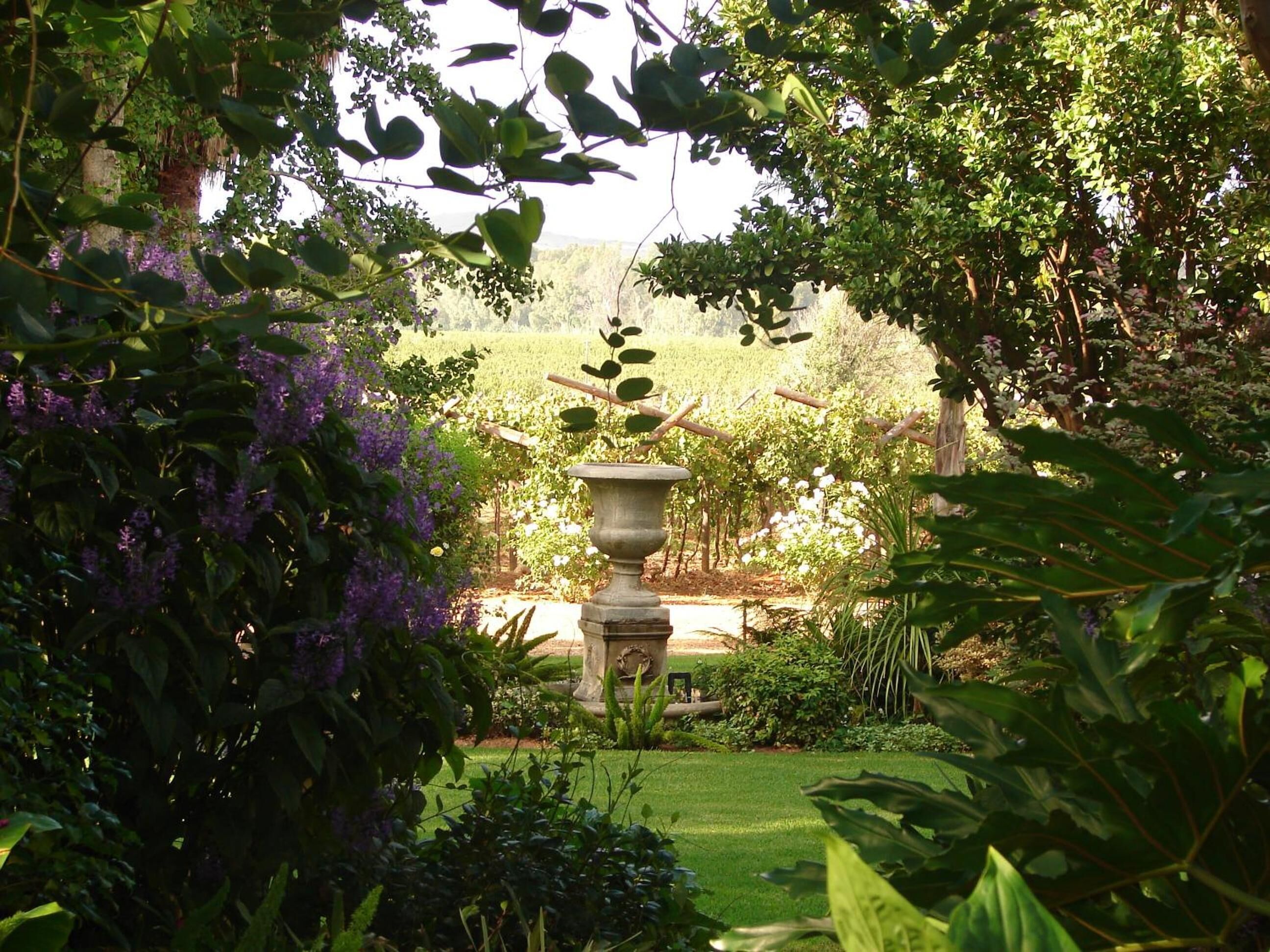 A garden with a central stone fountain, surrounded by lush greenery and flowering plants.