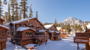 A row of wooden cabins with snow-covered roofs, set against a backdrop of a snow-covered mountain and pine trees.