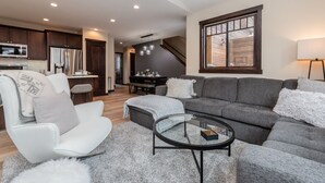 A modern living room with a gray sectional sofa, a white armchair, and a glass coffee table. A kitchen with dark wooden cabinets and stainless steel appliances is visible in the background.