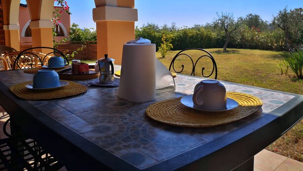 Une table à manger installée à l’extérieur, avec vue sur un jardin, une machine à café et des tasses bleues.