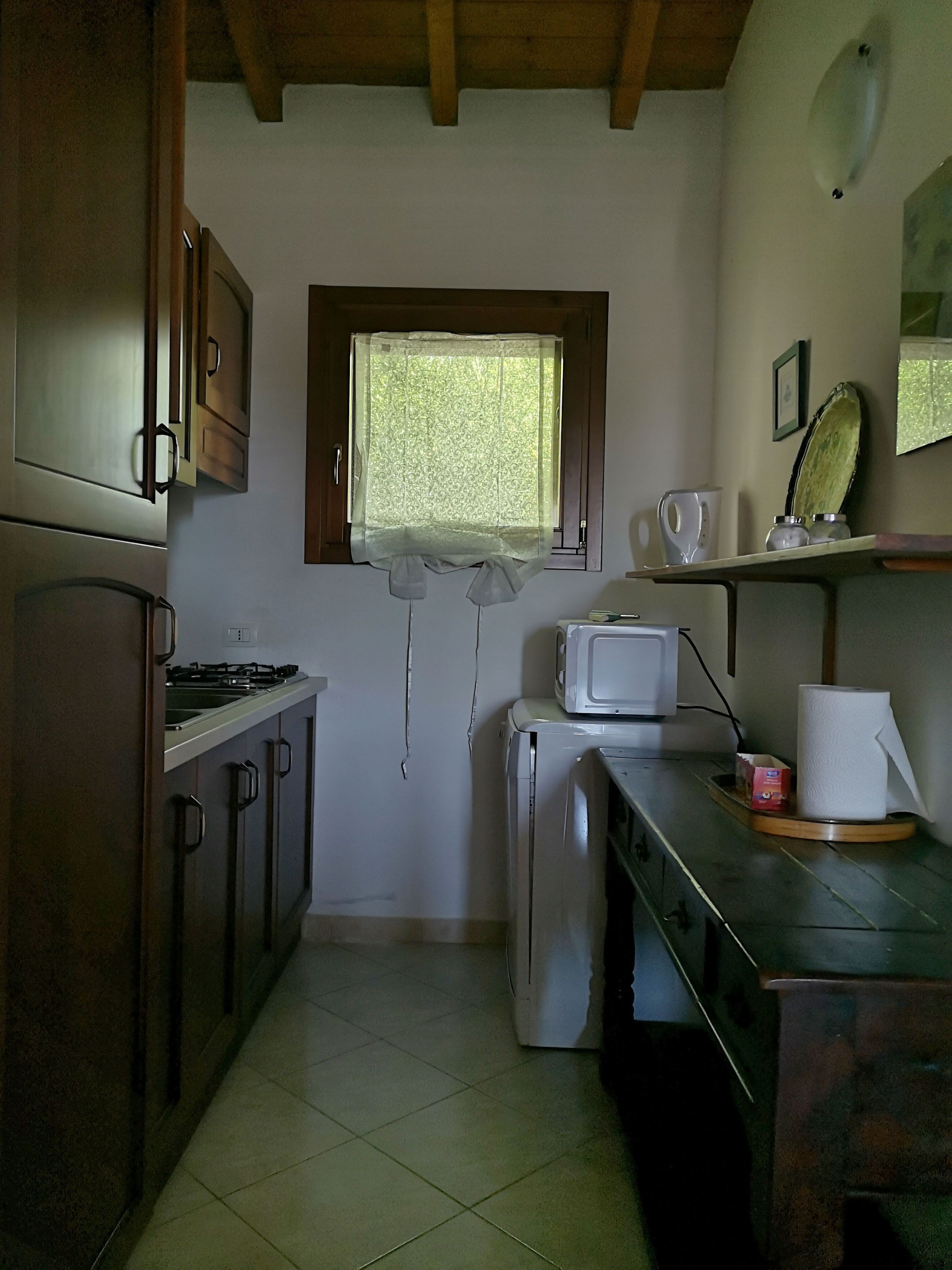 A compact kitchen with wooden cabinets, a microwave, a small refrigerator, and a window with a curtain.