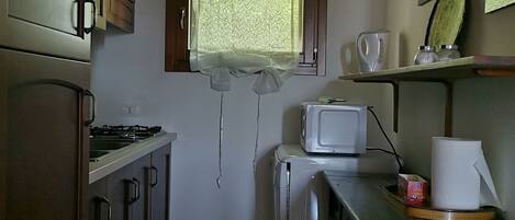 A compact kitchen with wooden cabinets, a microwave, a small refrigerator, and a window with a curtain.
