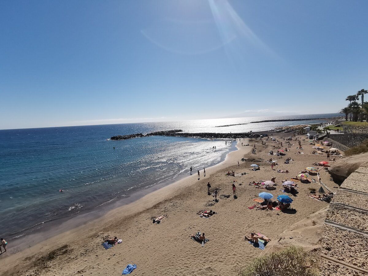 Una playa con gente tomando el sol y nadando, un muelle que se adentra en el agua, y un cielo despejado.