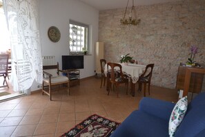A living room with a TV, a dining table with chairs, a blue sofa, and a stone wall.