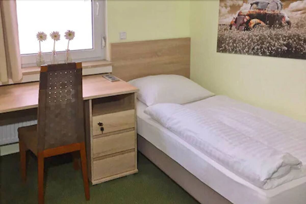 A bedroom with a single bed, a wooden desk with a chair, a window with flowers, and a wall-mounted picture.