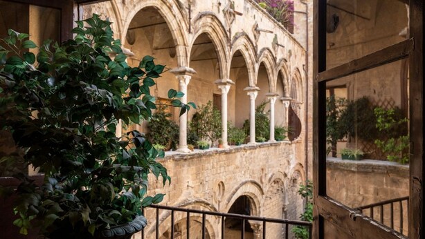 A stone building with arched windows and columns, viewed from a balcony with a potted plant. - Ajutamicristo by DomuSicily (Palermo)