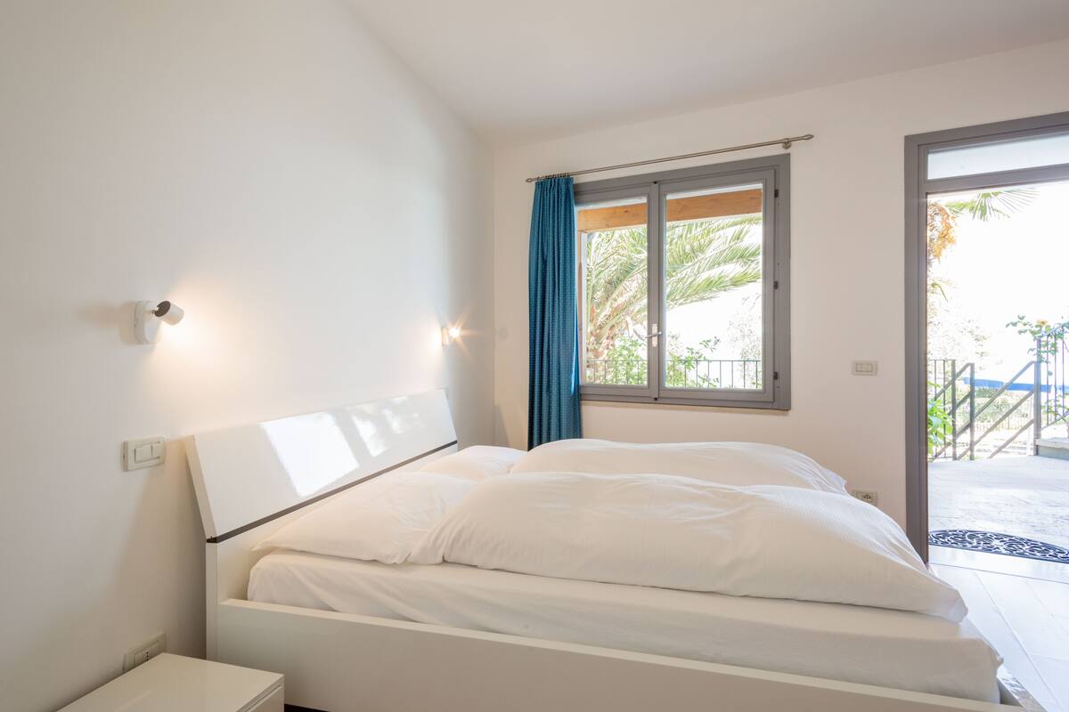 Un dormitorio con cama, mesita de noche, ventana con cortinas y vistas al exterior.