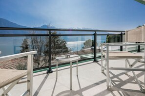 A balcony with a glass railing overlooking a lake and mountains. There are two white tables and a wicker chair. - Relax Hotel La Gardenia & Villa Oleandra (Limone sul Garda)