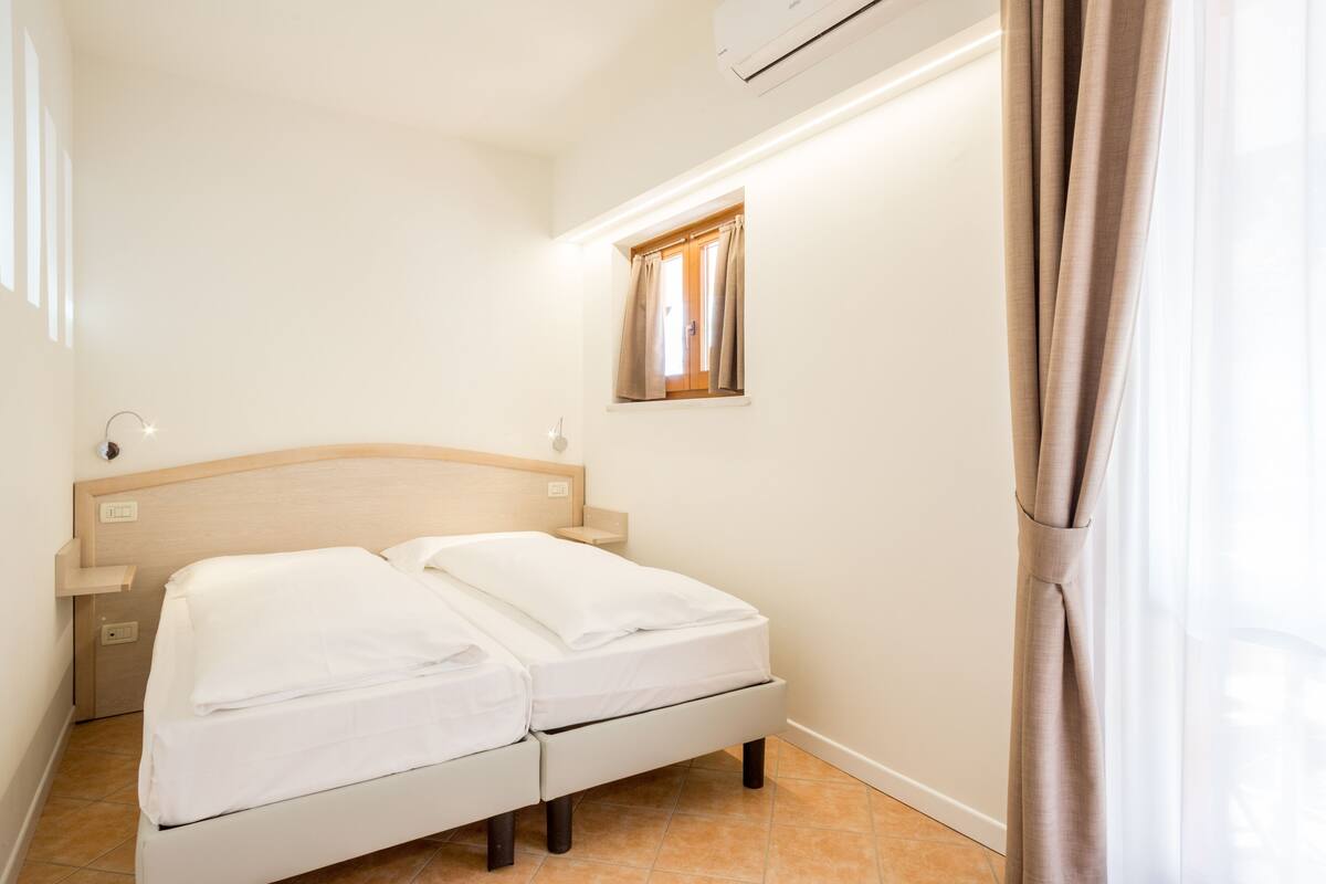 Una cama con dos camas individuales bien hechas, un cabecero de madera, una ventana pequeña con cortinas y un equipo de aire acondicionado arriba.