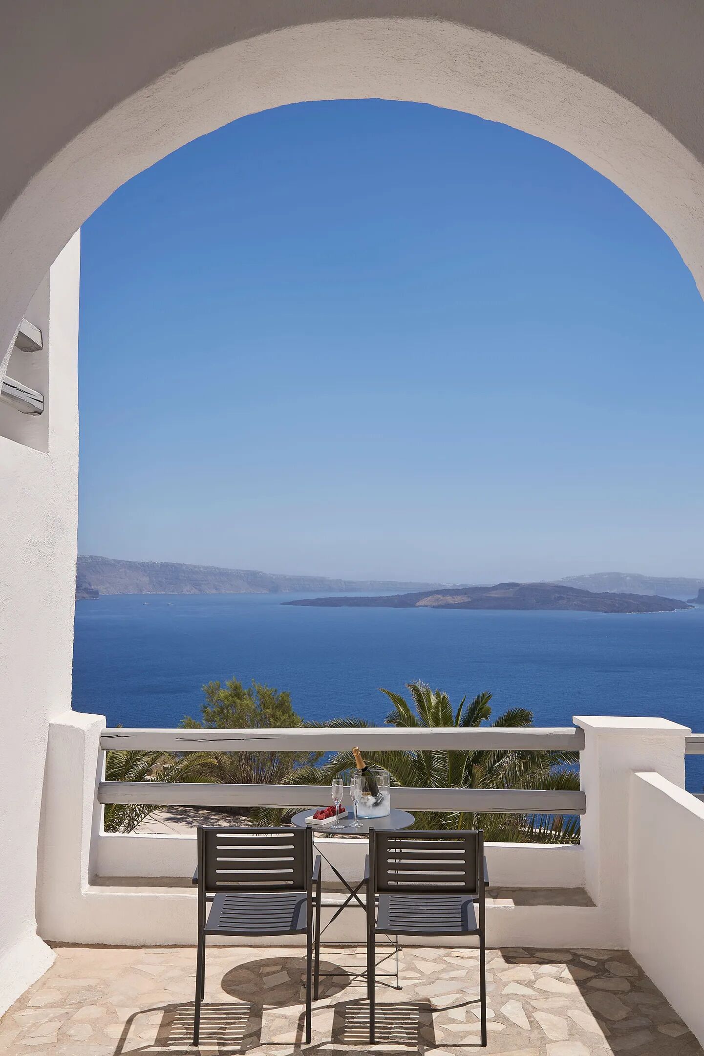 A balcony with two chairs and a table, overlooking a clear blue sea and distant land.