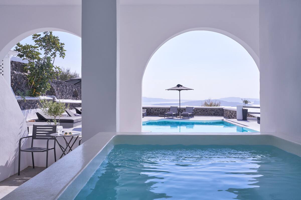 A pool area with clear blue water, a table with chairs, and a view of the landscape through an arched window.