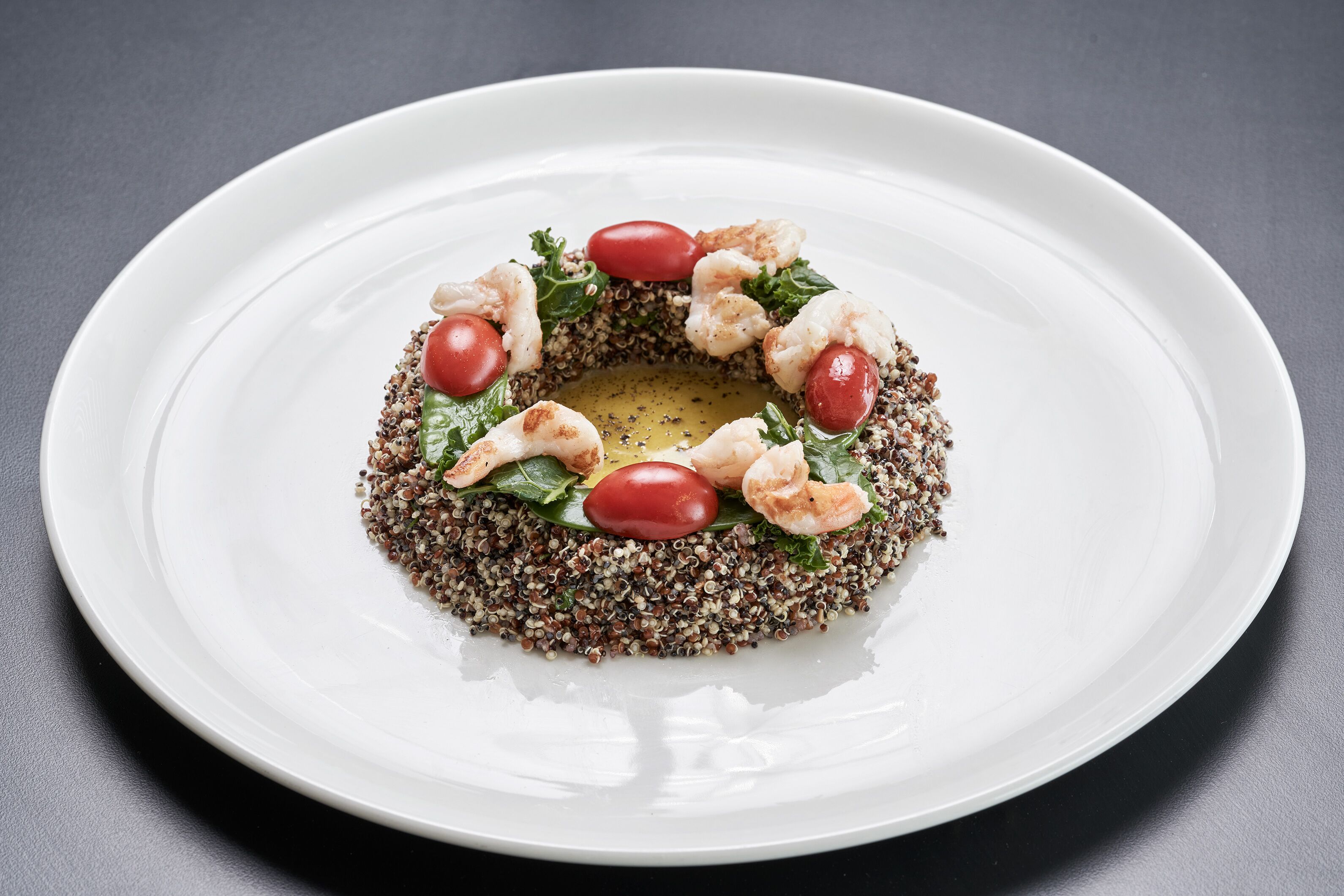 Un plato blanco con ensalada de quinoa, adornada con tomates cherry y camarones, sobre una superficie oscura.