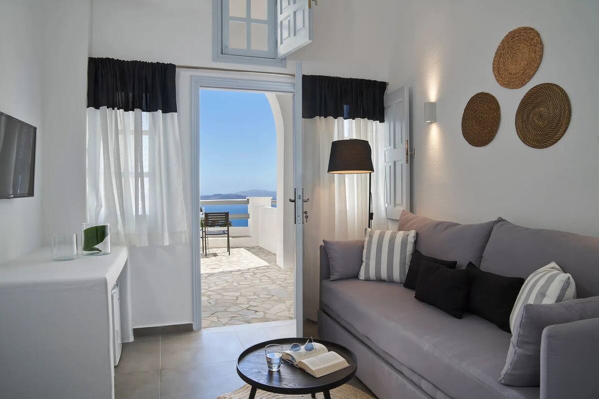 A modern living room with a grey sofa, a small coffee table with a book and glasses, and a view of the sea through a glass door.