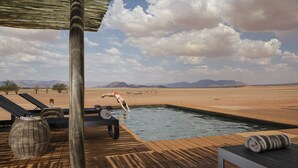 A pool area with lounge chairs, a wooden deck, and a desert landscape in the background. -  Kwessi Dunes (Sesriem)