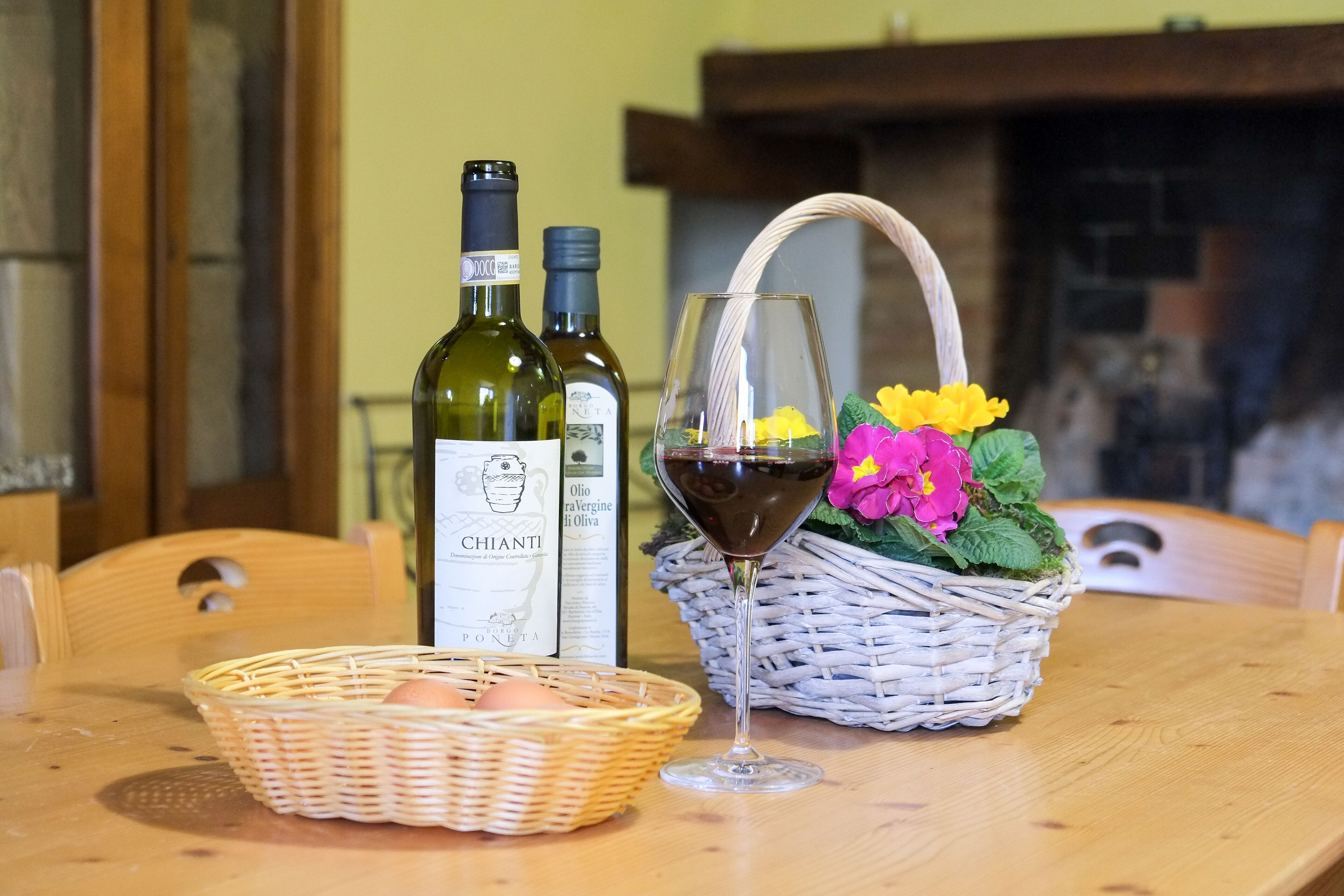 A wooden table with two wine bottles, a wine glass, a basket with eggs, and a basket with flowers.