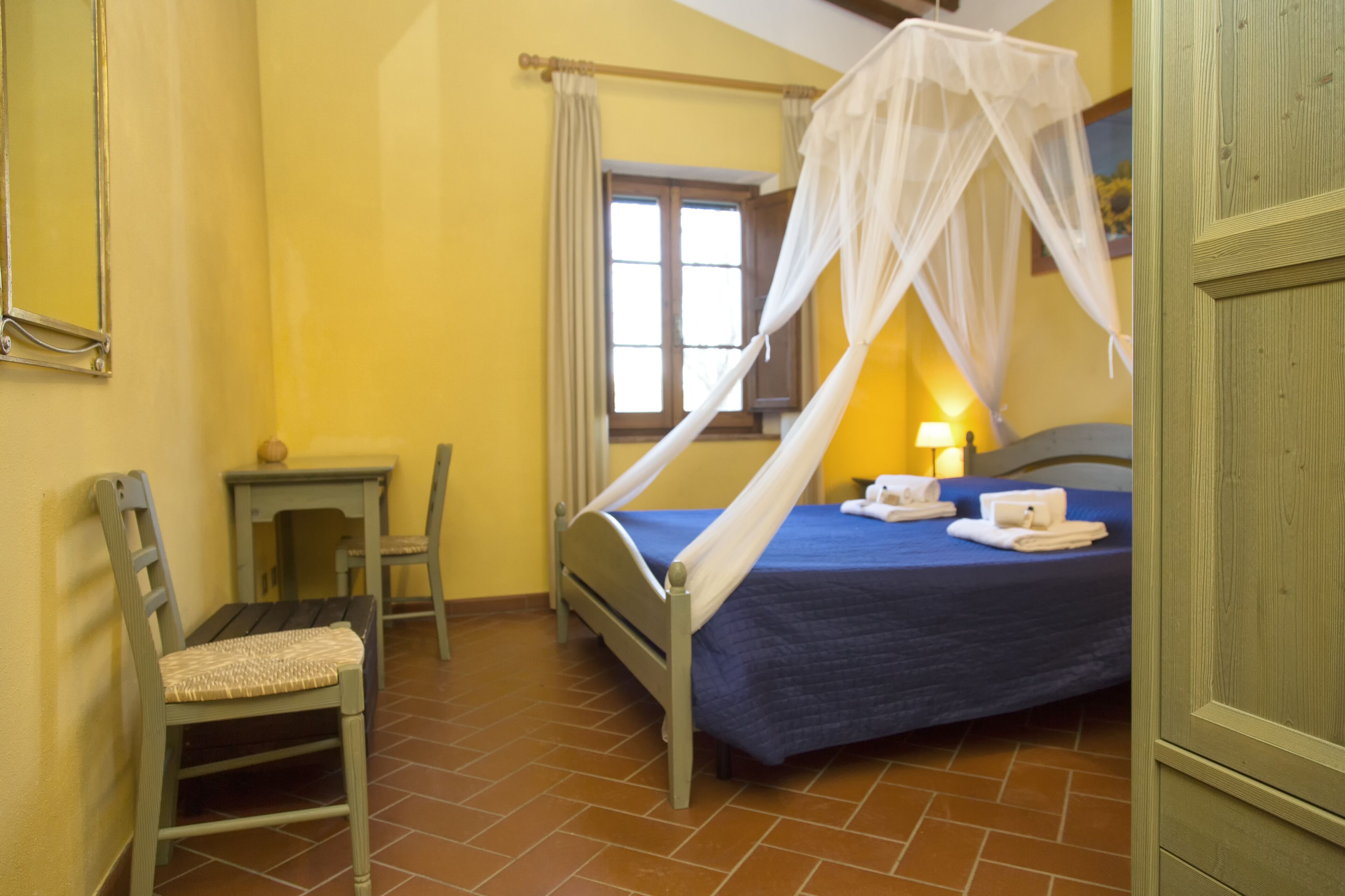 A bedroom with a bed, a chair, a nightstand, a window with curtains, and a bedside lamp.