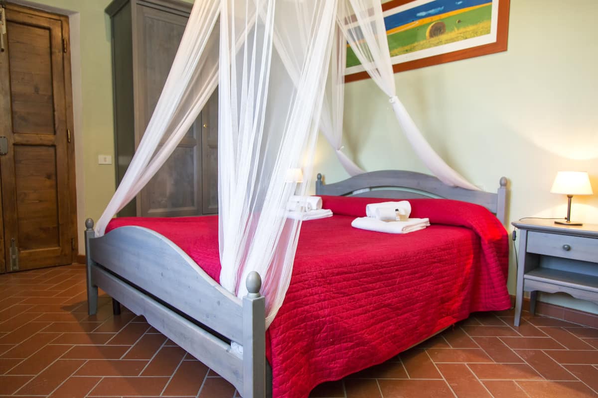 A bed with a red bedspread and white canopy, a bedside table with a lamp, and a wooden door in the background.