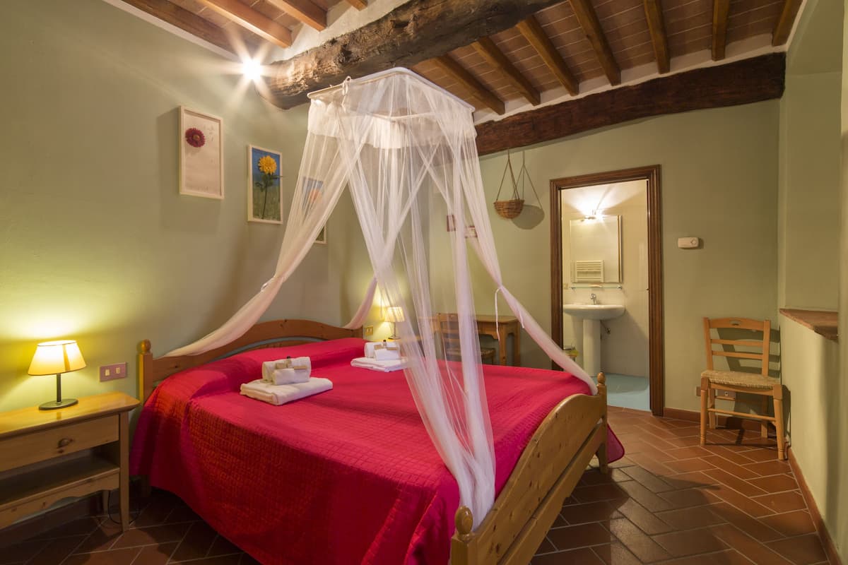 A bedroom with a four-poster bed, a bedside table with a lamp, a chair, and a bathroom visible through an open door.