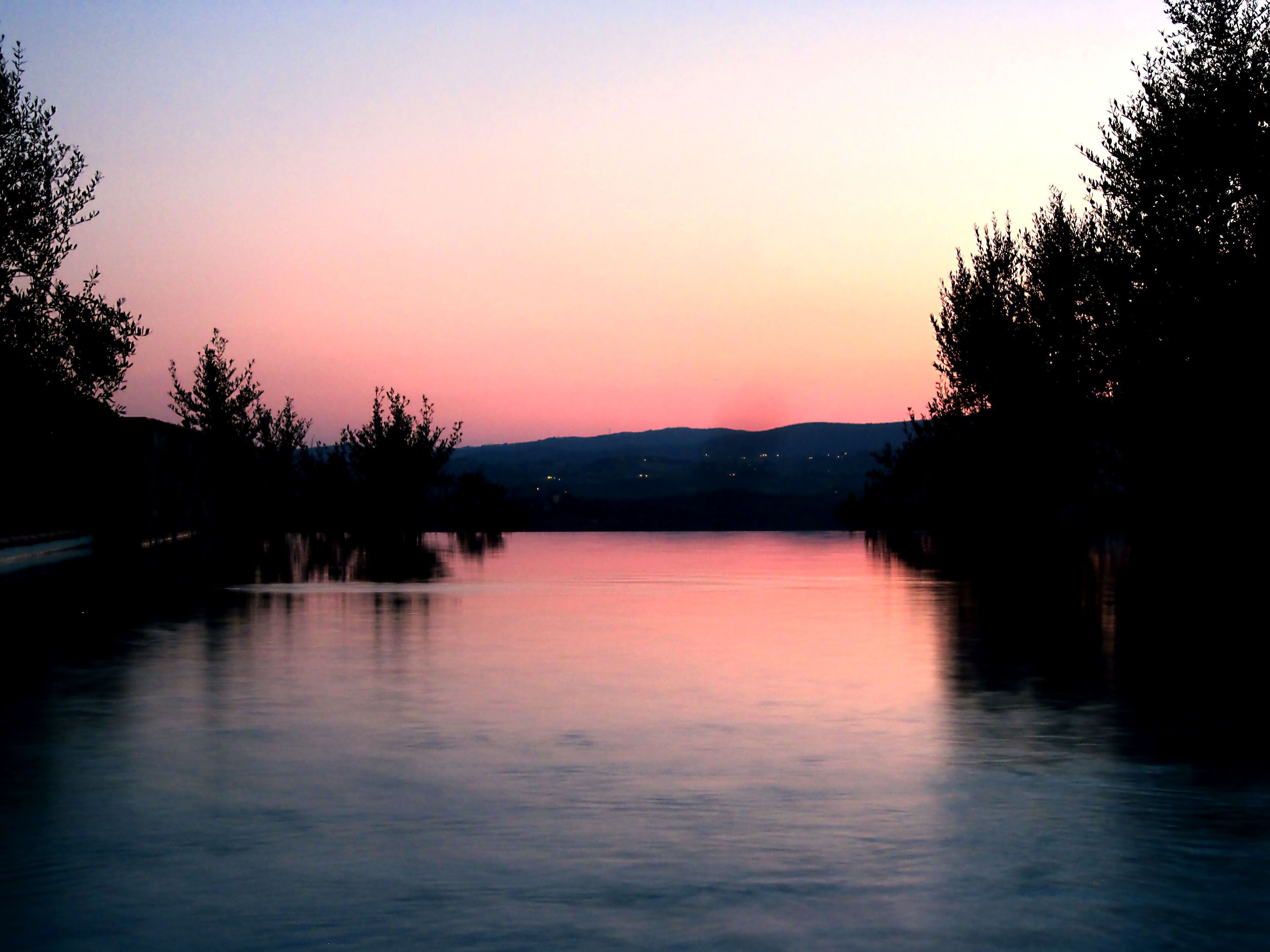 A calm lake at dusk with trees on either side and distant lights on the horizon.