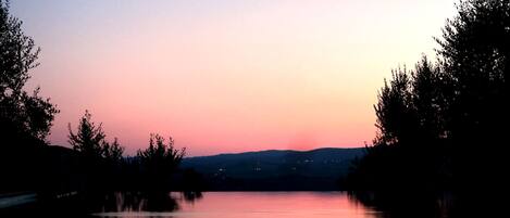A calm lake at dusk with trees on either side and distant lights on the horizon.