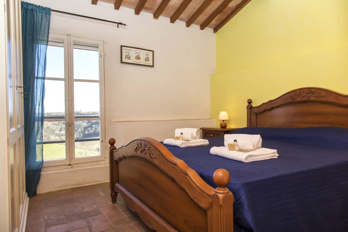 A bedroom with a wooden bed, a window with blue curtains, a bedside table with a lamp, and a view of the outdoors.