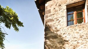 Un edificio de piedra con puerta y ventana de madera, rodeado de follaje y plantas en macetas.