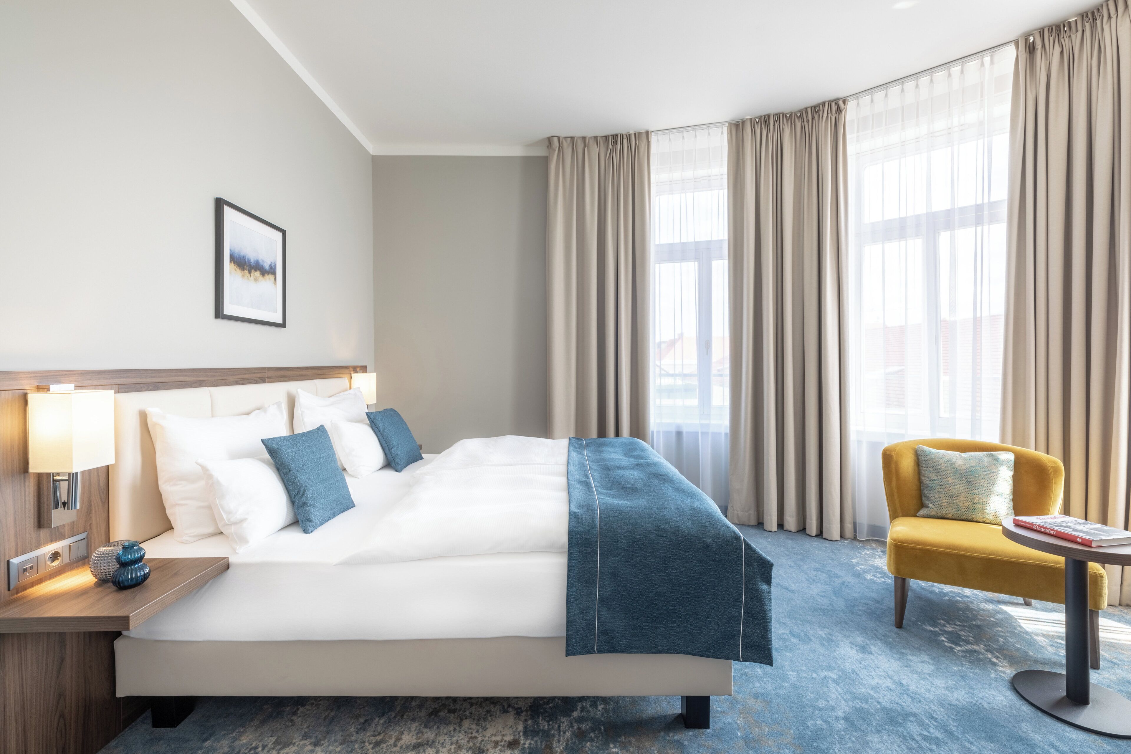 A hotel room with a large bed, a bedside table with a lamp, a chair, and a window with curtains.