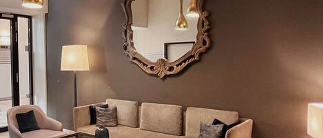 A modern living room with a beige sofa, two armchairs, a glass coffee table, and a large ornate mirror.
