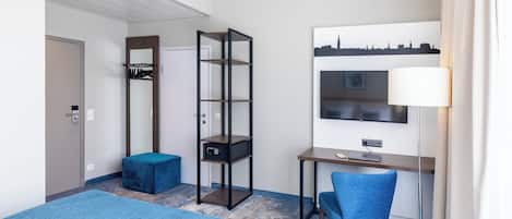 A hotel room with a bed, a desk with a chair, a wardrobe, a mirror, a shelf unit, and a cityscape artwork.