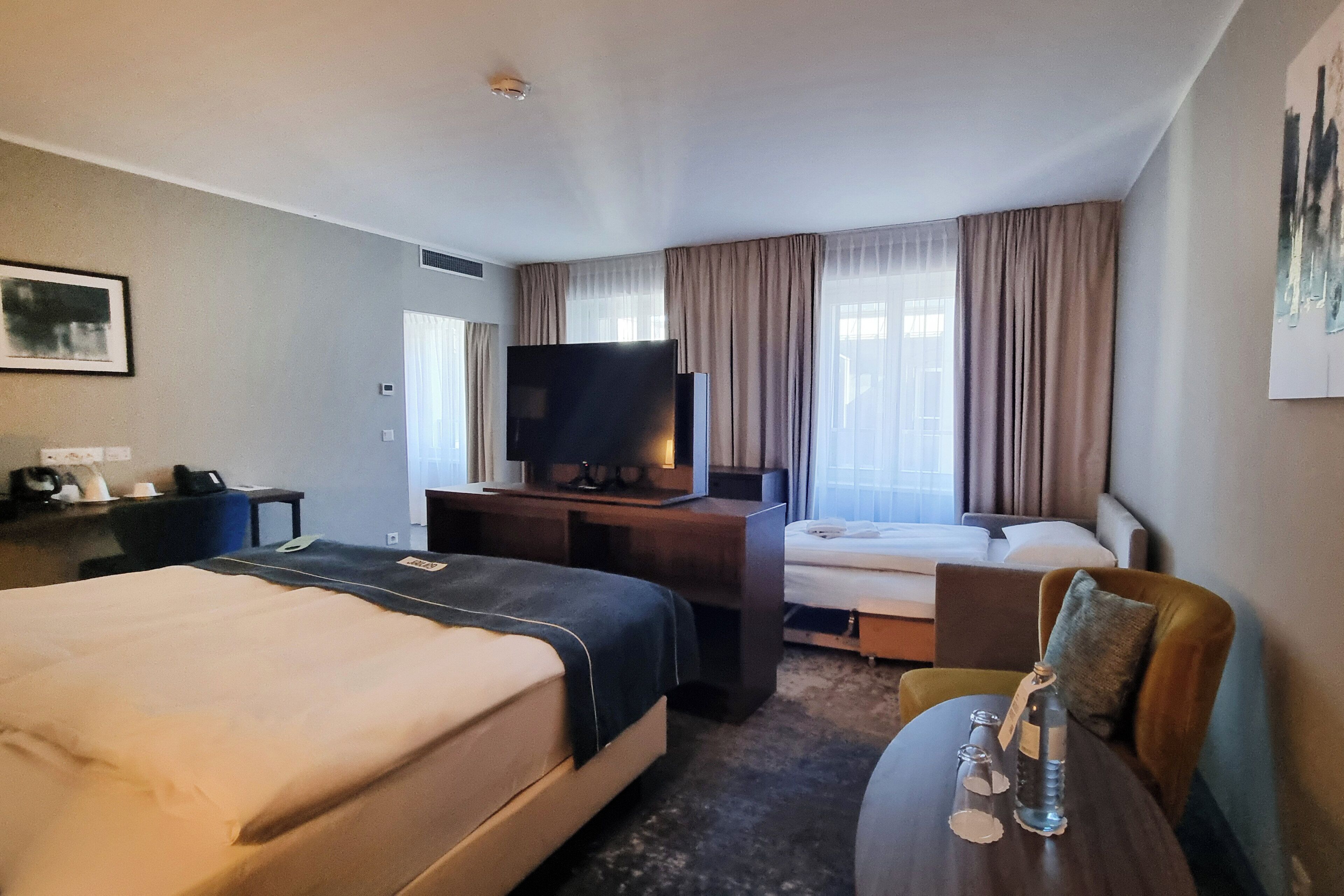 A hotel room with two beds, a desk with a television, a chair, and a small table with a bottle and glasses.