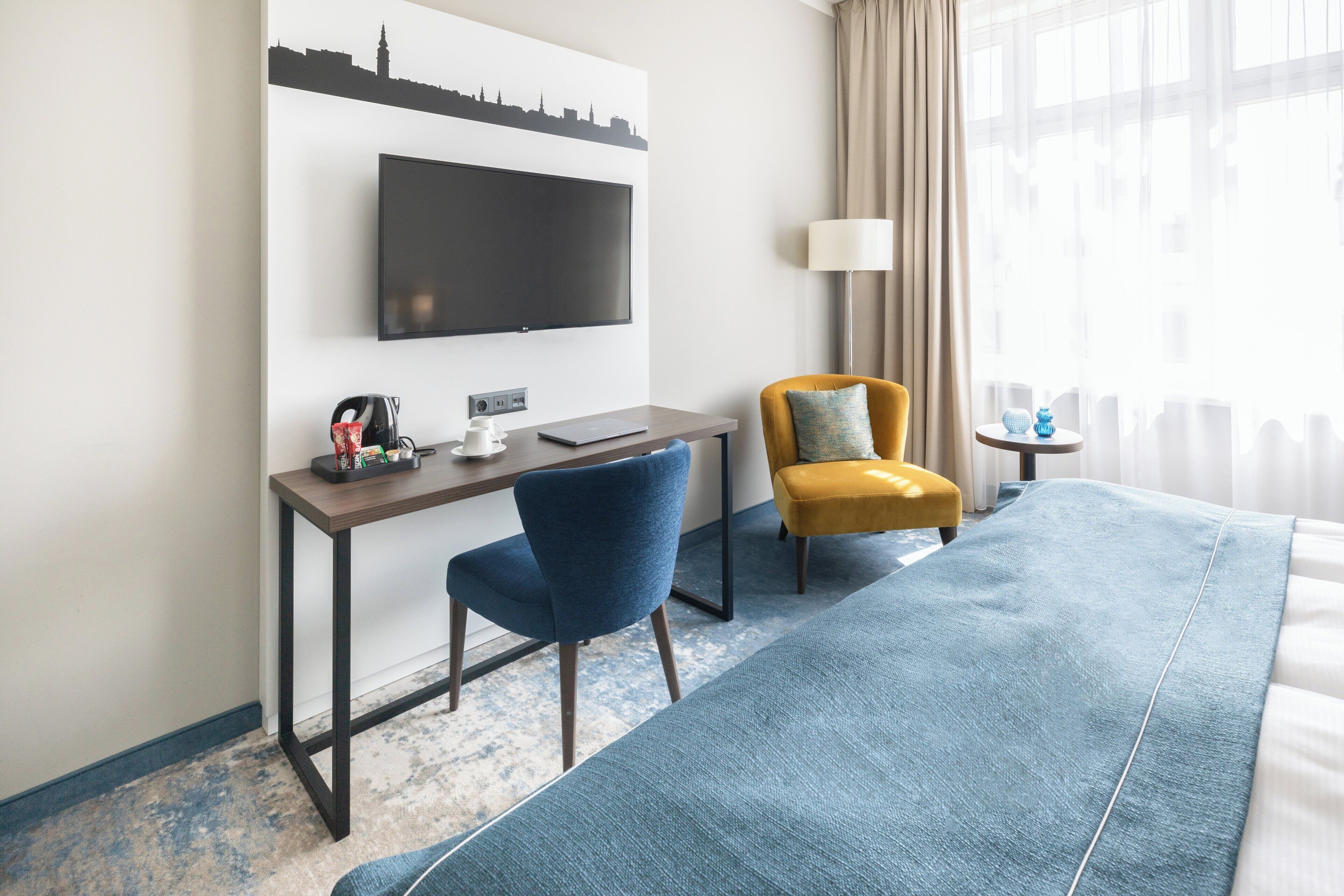 A hotel room with a large bed, a desk with a laptop, a TV mounted on the wall, and a city skyline artwork.