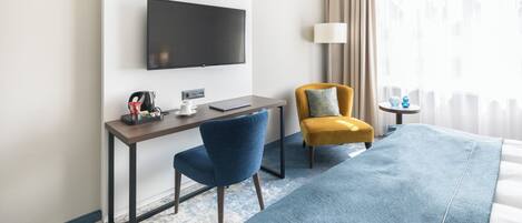 A hotel room with a large bed, a desk with a laptop, a TV mounted on the wall, and a city skyline artwork.