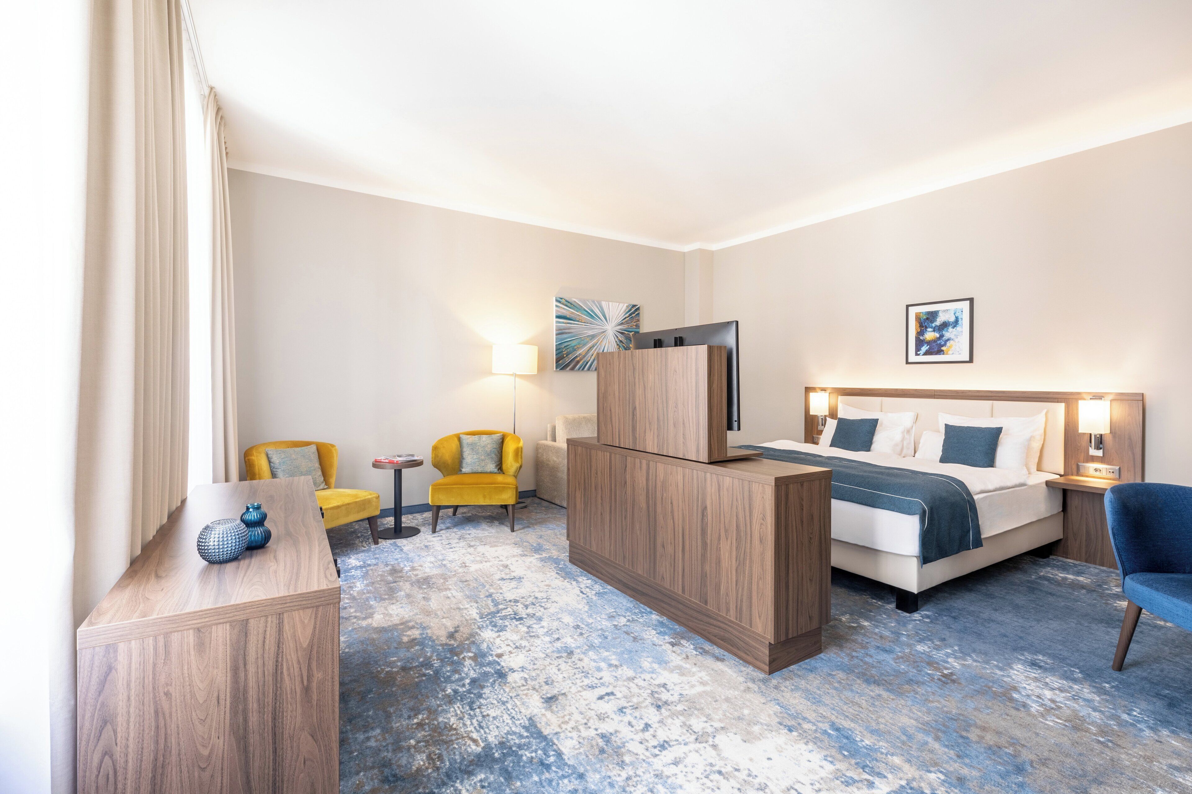 A hotel room with a bed, a desk, a chair, a lamp, and artwork on the walls.