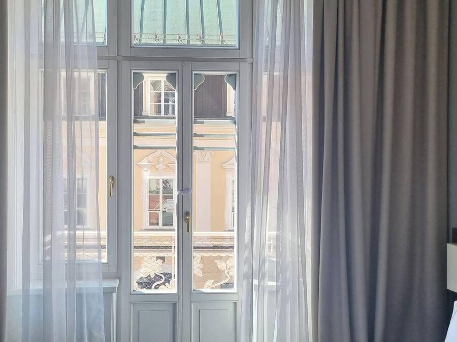 A room with a large window, sheer curtains, and a view of a building outside.