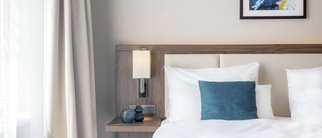 Una cama bien hecha con ropa de cama blanca y azul, una lámpara de noche y un pequeño jarrón decorativo sobre una mesita de noche de madera.
