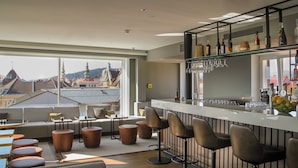 A modern bar area with a view of the cityscape, featuring a bar counter, stools, and a variety of bottles and glasses. - Garner Hotel Klagenfurt Moser Verdino by IHG (Klagenfurt am Woerthersee)